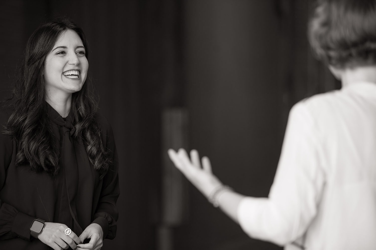 The image shows Nicole Padua, who is smiling and engaged in a conversation. She has long dark hair and is wearing a dark blouse with a watch on her left wrist. Her expression reflects joy and interest as she listens to the person in front of her, who is partially visible and gesturing with their hands. The image is in black and white, giving it a timeless, professional feel.